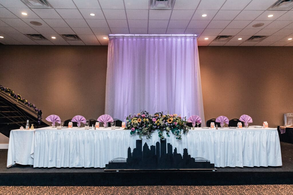 Wedding reception head table with purple uplighting, white linens, and city skyline backdrop at The Fez