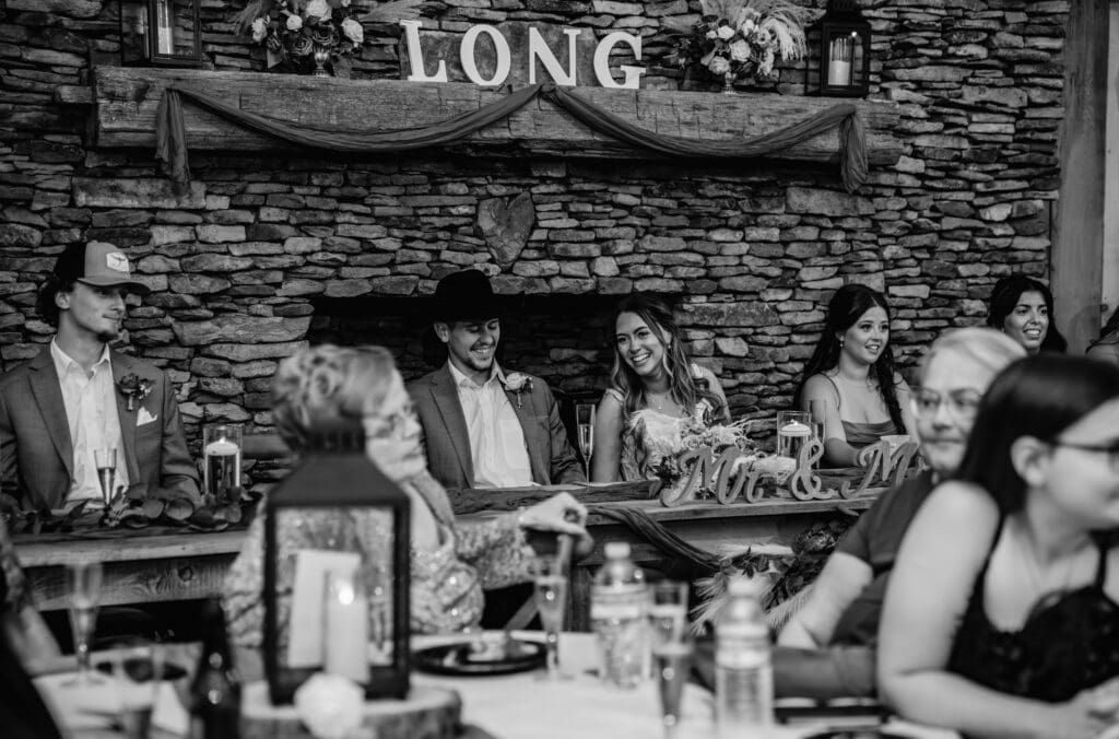 Wedding couple laughs and smiles during speechs in rustic venue with warm lighting at Hinckston Run Farm