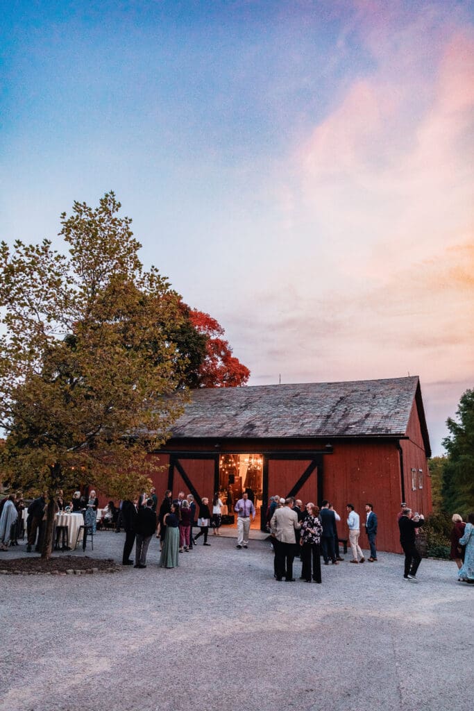 Wedding guests talking and mingling with Pinehall reception barn in the background at sunset