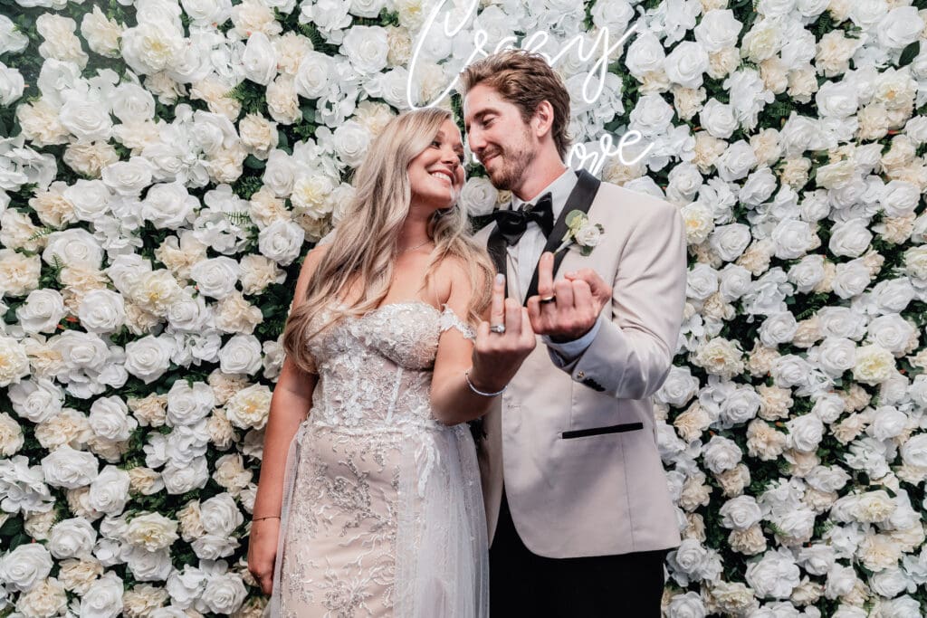 Bride and groom show off their wedding rings and pose in front of a floral wall with a neon sign at their Le Mont micro wedding reception in Pittsburgh