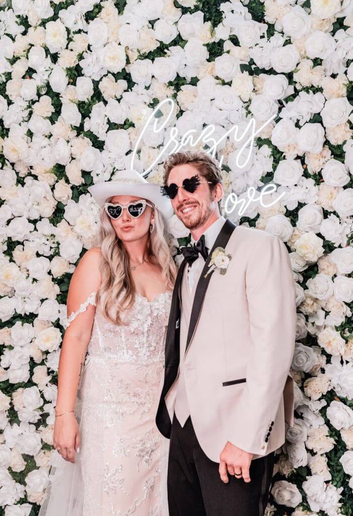 Bride and groom wearing fun sunglasses and posing in front of a flower wall with neon sign at Le Mont