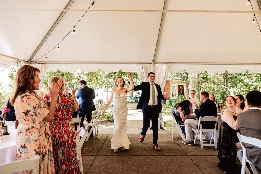 Wedding guests cheering as newlyweds walk through a tented reception area at Schenley Park Welcome Center