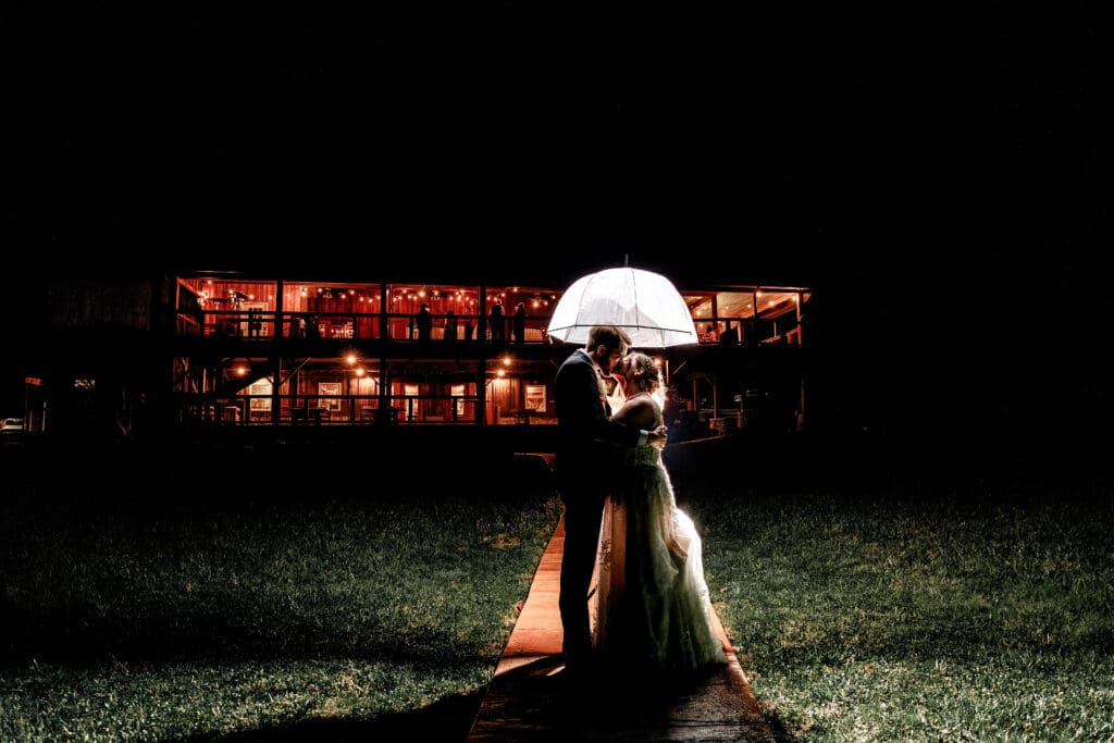 Romantic nighttime wedding photo of bride and groom under an umbrella outside the glowing barn venue at Lingrow Farm