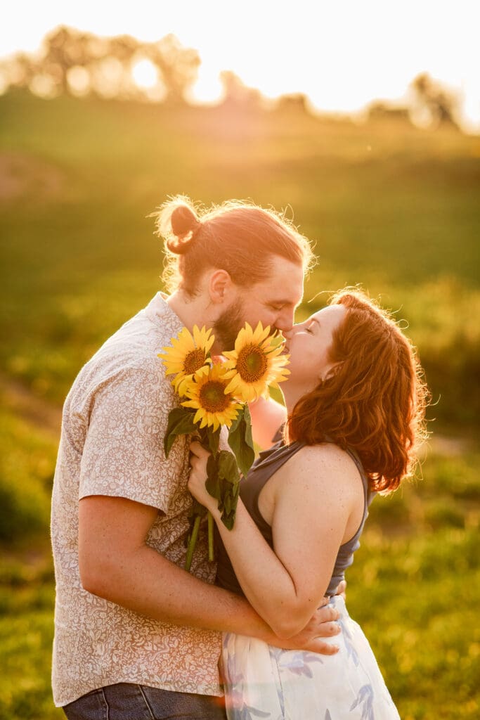 Engaged couple embracing in sunlit meadow during golden hour with dreamy warm light at Schwirian Sunflower Festival