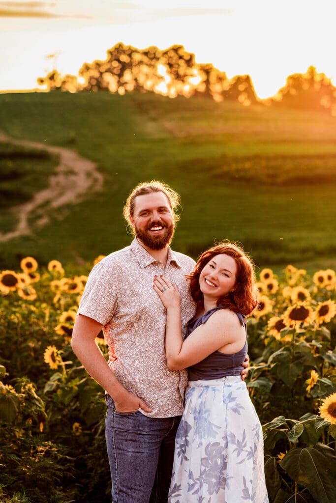 Engaged couple standing together in a golden sunflower field at sunset with rolling hills at Schwirian Sunflower Festival