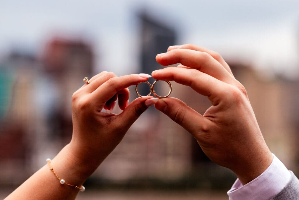 Hands holding wedding rings against a blurred Pittsburgh cityscape at Sheraton Station Square