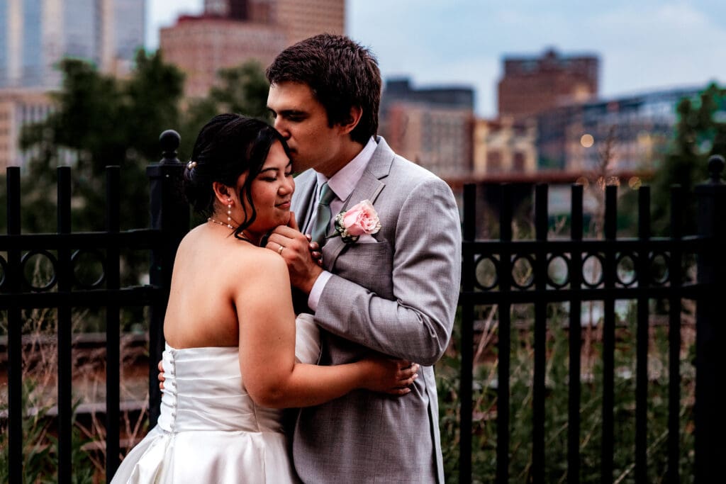 Wedding couple embraces in front of a black metal fence with Pittsburgh skyline near Sheraton Station Square