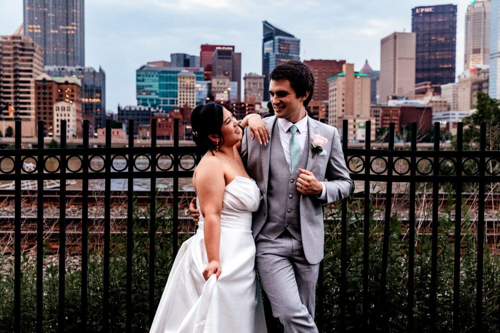 Wedding couple poses against a black iron fence with Pittsburgh skyline at Sheraton Station Square