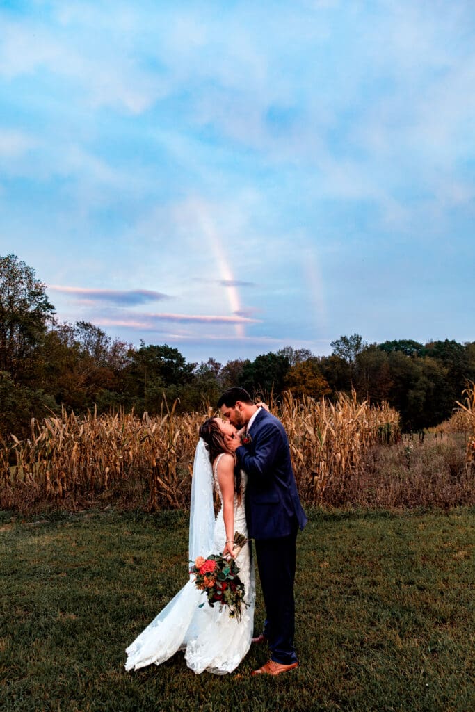 Wedding couple embraces beside a cornfield under rainbow skies at Hinckston Run Farm