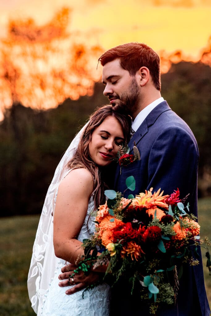 Couple embraces at sunset holding vibrant red and orange bouquet at Hinckston Run Farm
