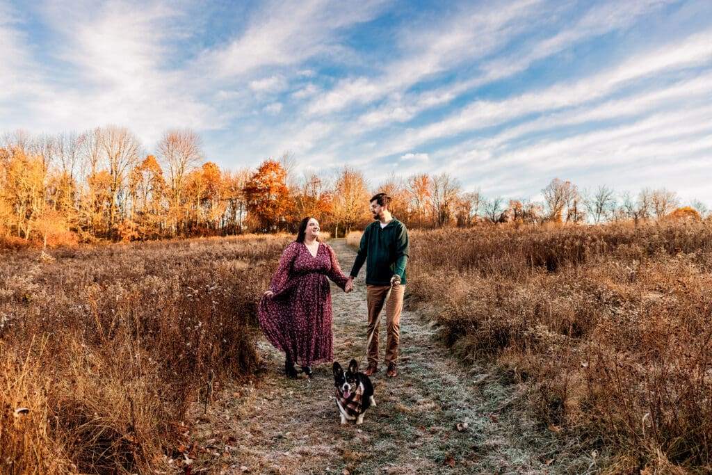 Couple walks together with their corgi through a field at Sewickley Heights Borough Park during an engagement photo session