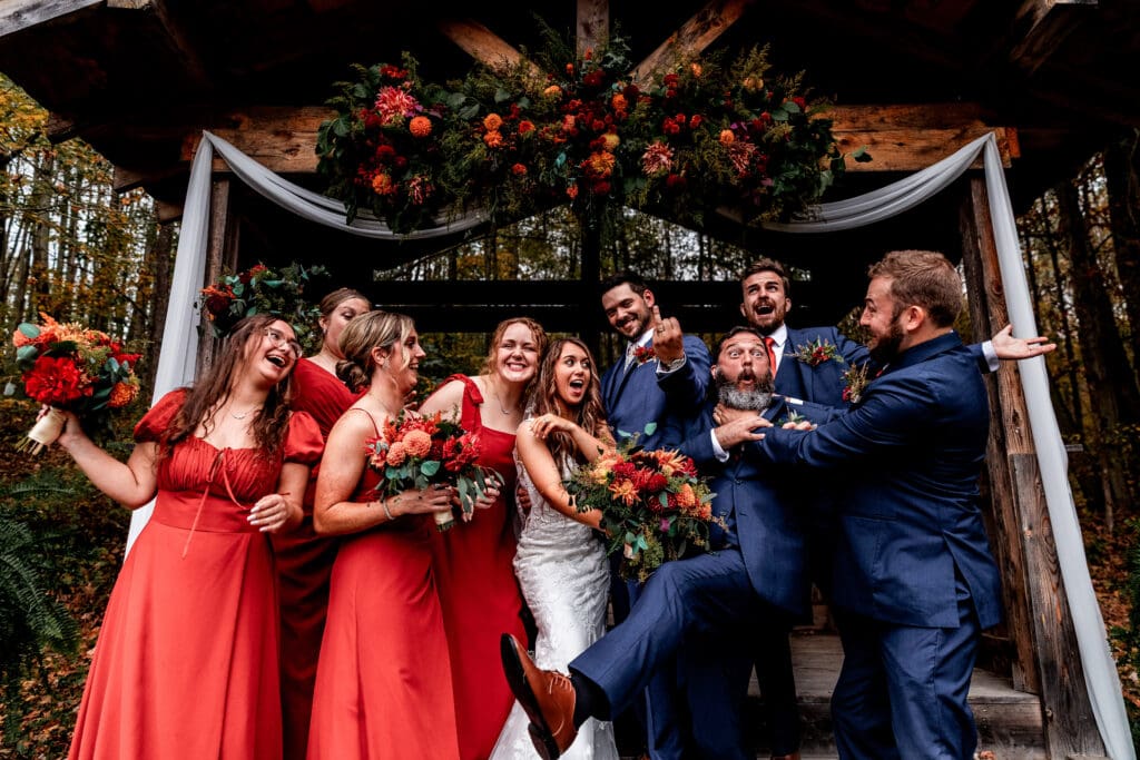 Wedding party in navy suits and red dresses poses playfully beneath ceremony arch at Hinckston Run Farm