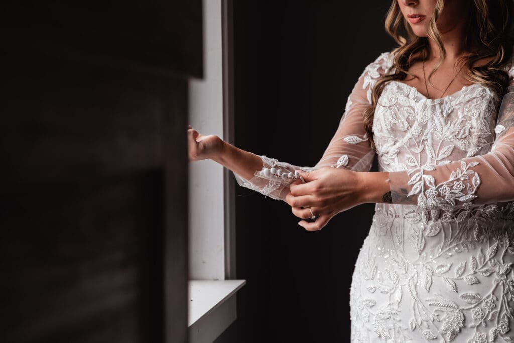 Bride buttoning lace sleeves by window during wedding morning at Hinckston Run Farm