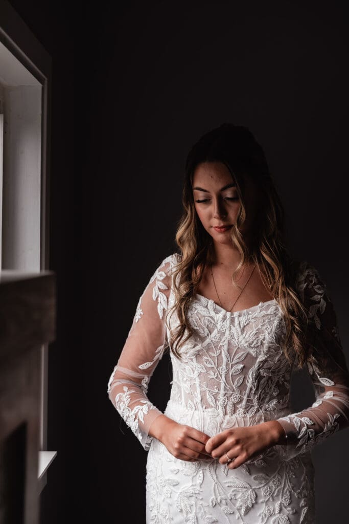 Bride in lace wedding dress standing by window in dramatic light at Hinckston Run Farm