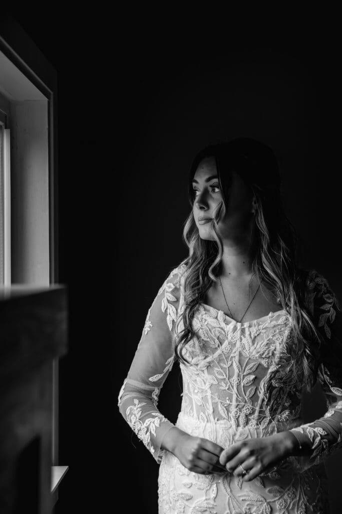 Black and white photo of bride looking out window while getting ready at Hinckston Run Farm