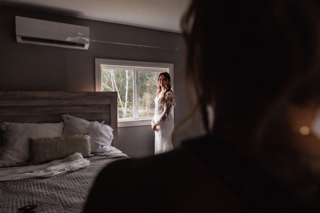 Photo over mother’s shoulder as she looks at bride in wedding dress by window at Hinckston Run Farm