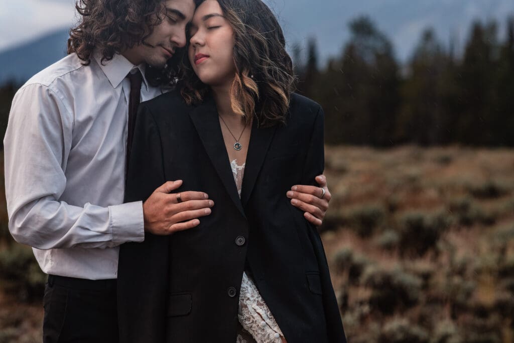 Close-up of an eloping couple’s hands wrapped together while they snuggle in front of the Teton mountains.