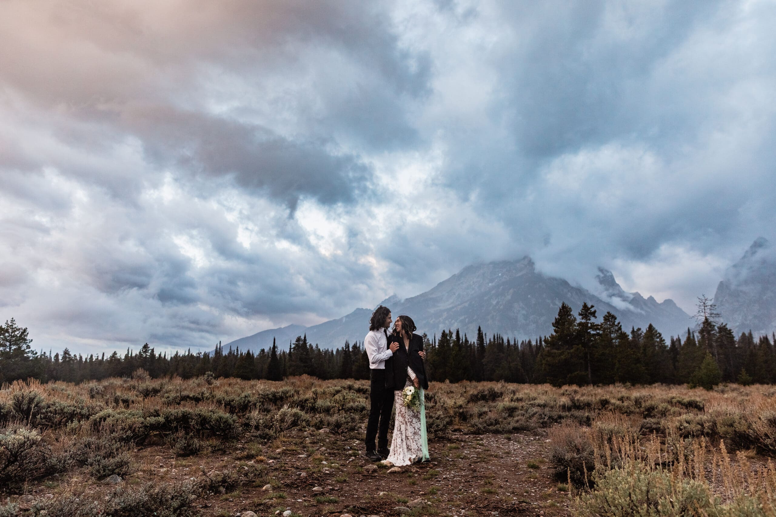 Couple standing close together in Grand Teton National Park with the bride wearing the groom’s jacket and the Teton peaks behind them.