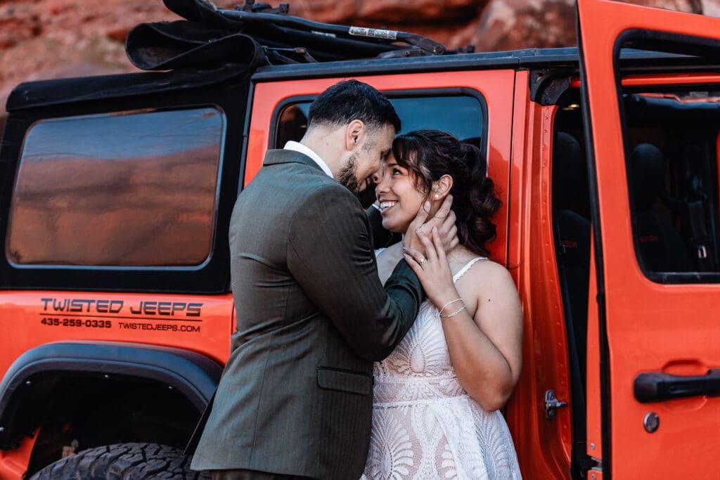 Couple leaning on a red Jeep during their Moab Utah elopement surrounded by red rock canyon scenery