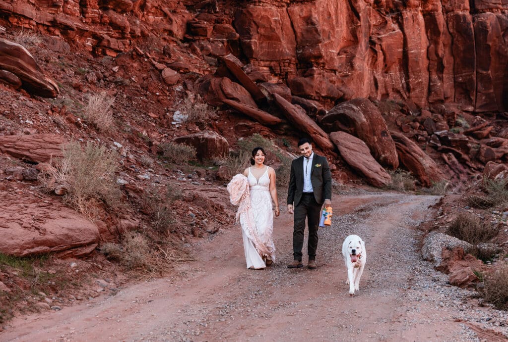 Eloping couple walking hand in hand with their dog across the red rock desert during their dog-friendly Moab elopement.