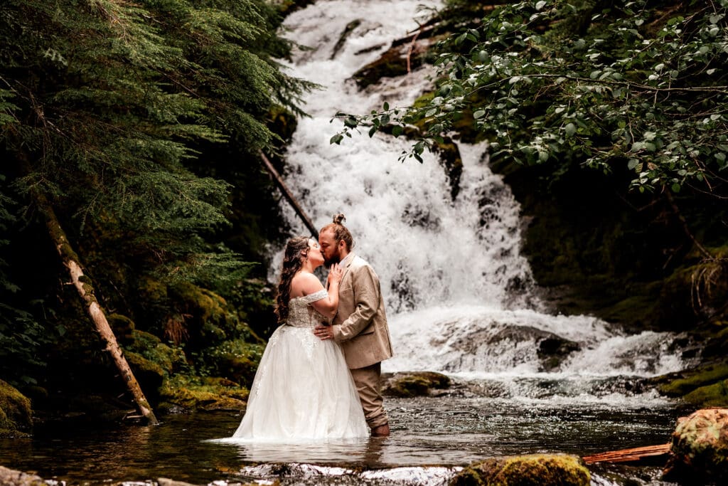 Eloping couple sharing a kiss while standing in the water with a waterfall flowing behind them in Mount Rainier National Park.
