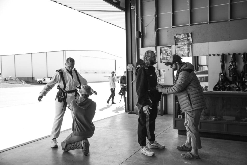 Couple smiling as a skydiving instructor secures her harness before a tandem skydive during the start of their adventurous elopement day.
