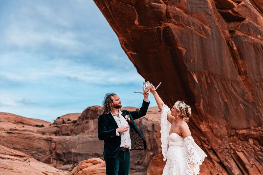 Just-married couple cheering and drinking champagne from champbongs after their elopement ceremony at Corona Arch near Moab, Utah.