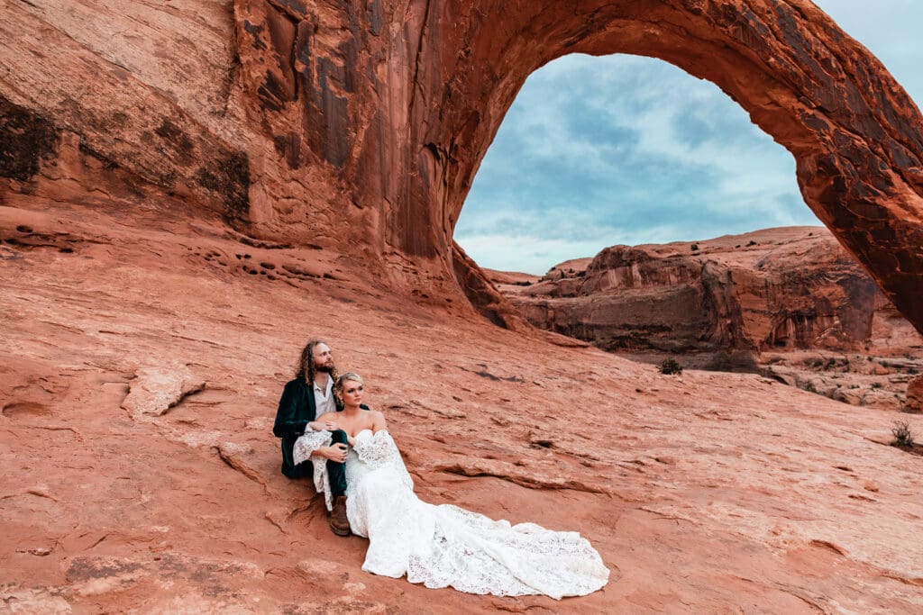 Newly married couple sitting together after exchanging vows with Corona Arch towering in the background during their Moab elopement.