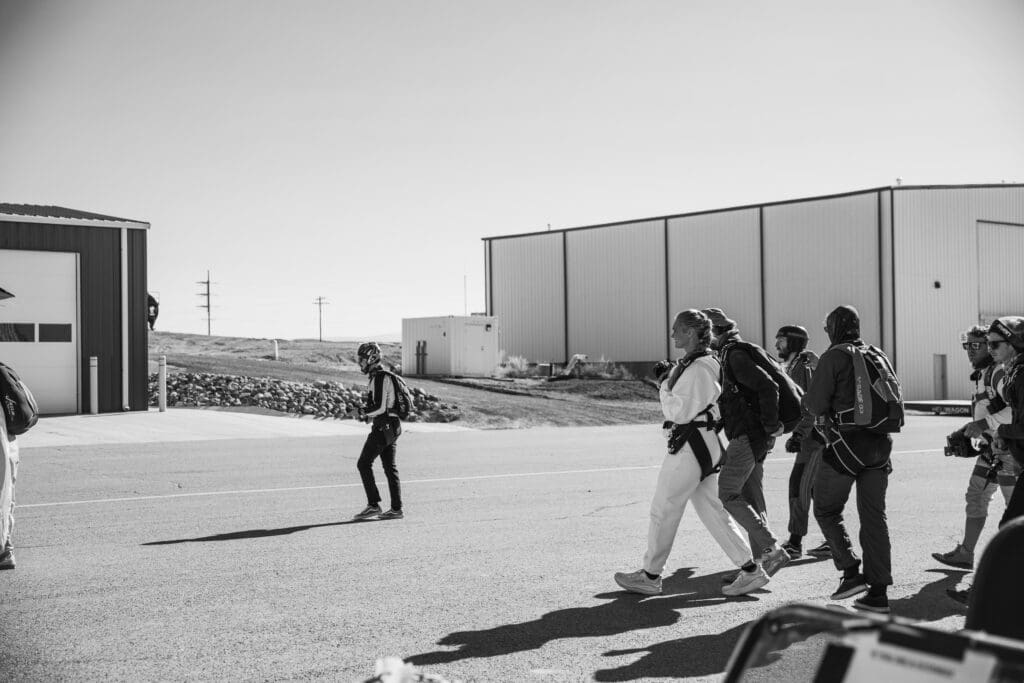 Eloping couple walking across the airfield toward the plane in their skydiving gear before their tandem jump adventure.