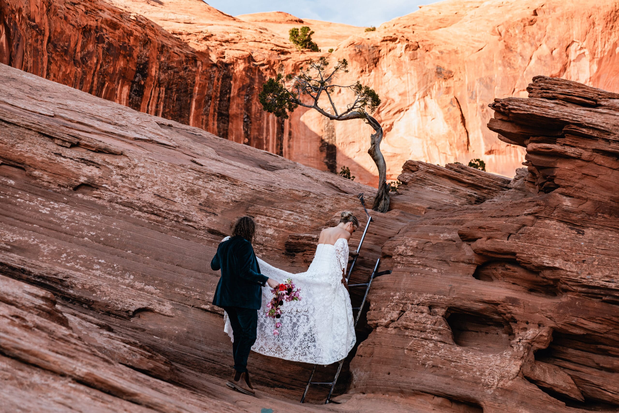 Groom holding the bride’s dress as she climbs a ladder along the Corona Arch trail during their adventurous Moab elopement.