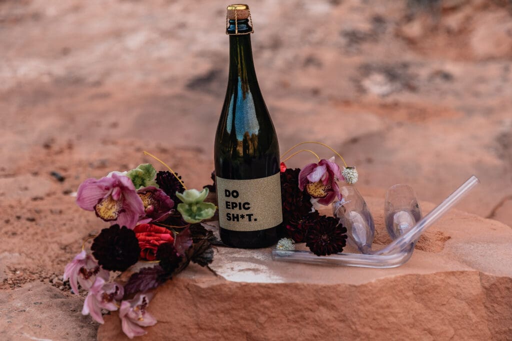 Flat lay of elopement details including champagne, flowers, vow book, and wedding rings photographed during a Corona Arch elopement in Moab, Utah.