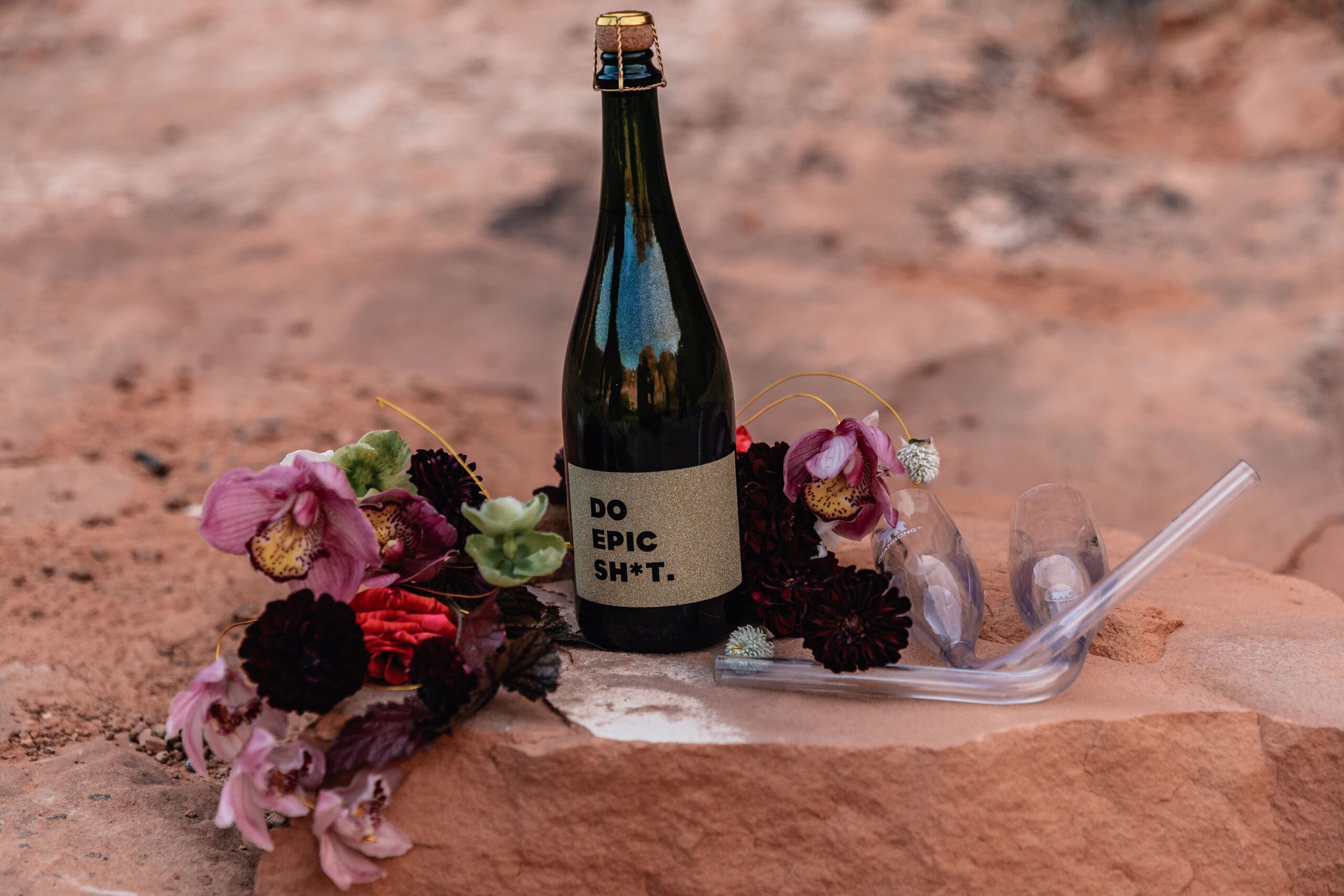Flat lay of elopement details including champagne, flowers, vow book, and wedding rings photographed during a Corona Arch elopement in Moab, Utah.