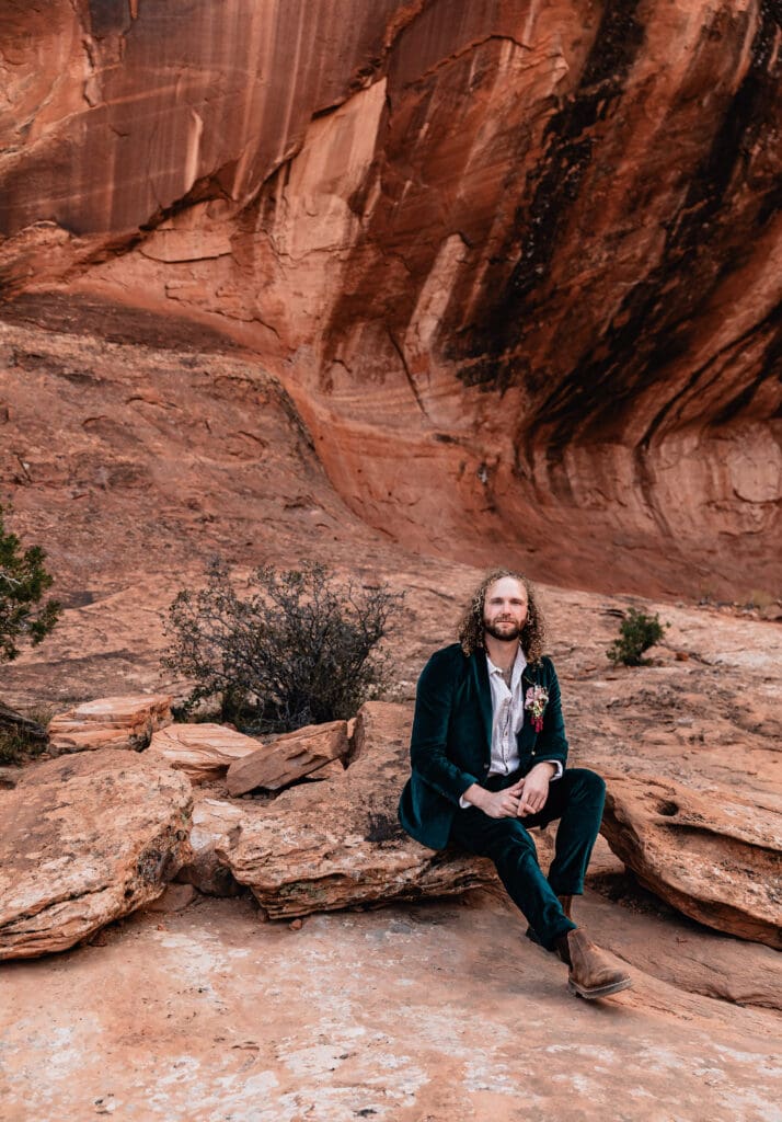 Groom sitting in a green suit on the red rock landscape while waiting for the ceremony to begin during a Corona Arch elopement in Moab, Utah.