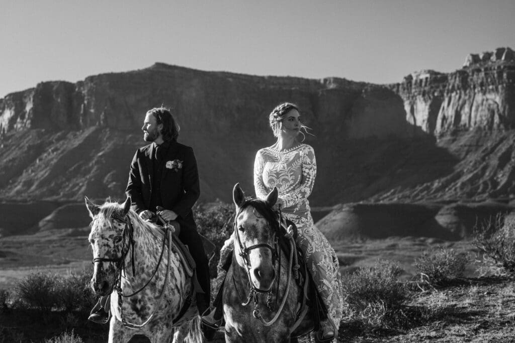 Black and white photo of an eloping couple riding horses side by side during the horseback riding portion of their adventure elopement.