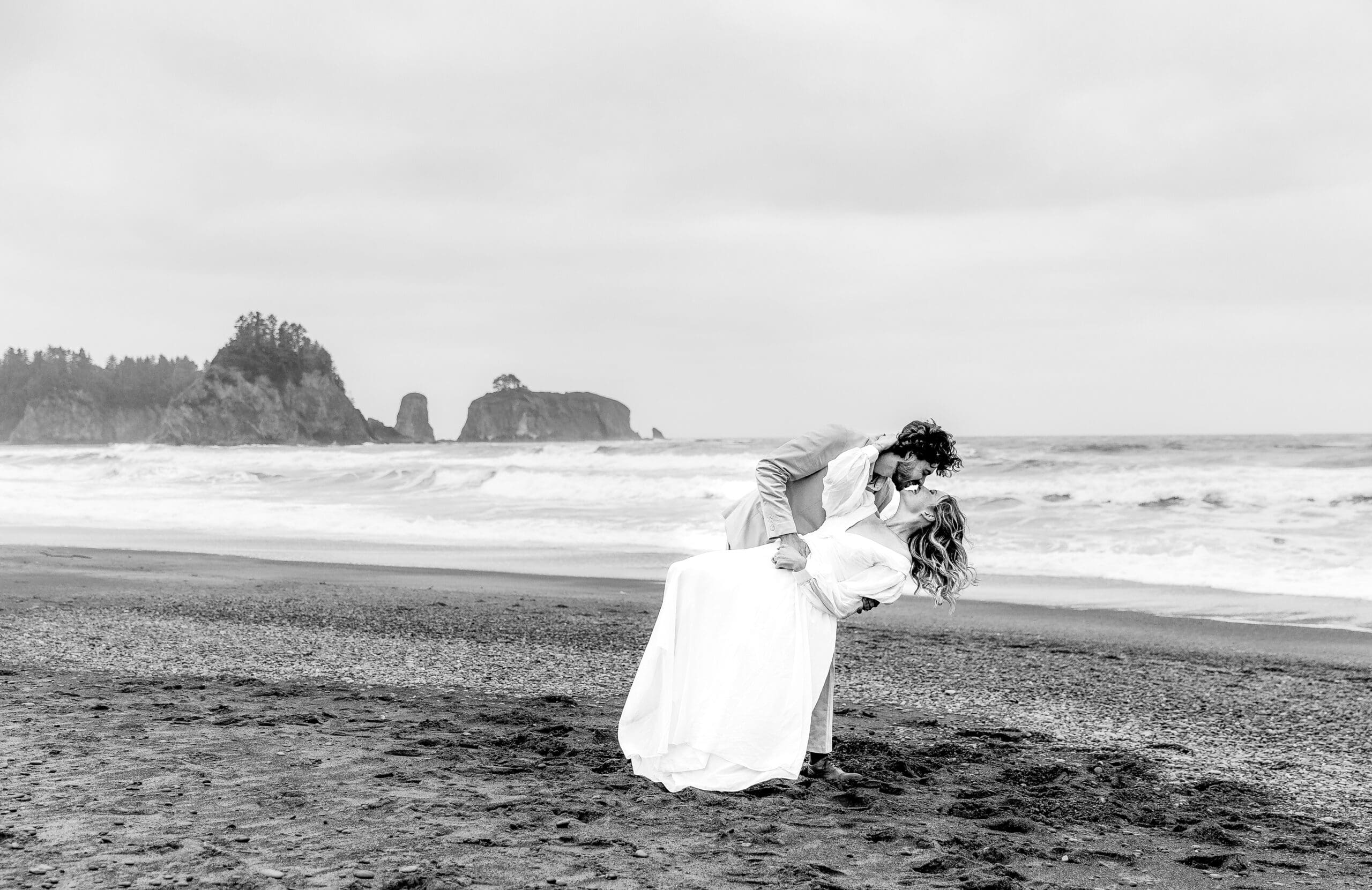 Black and white photo of a couple sharing a dip kiss on the rugged shoreline of Rialto Beach during their Washington elopement.