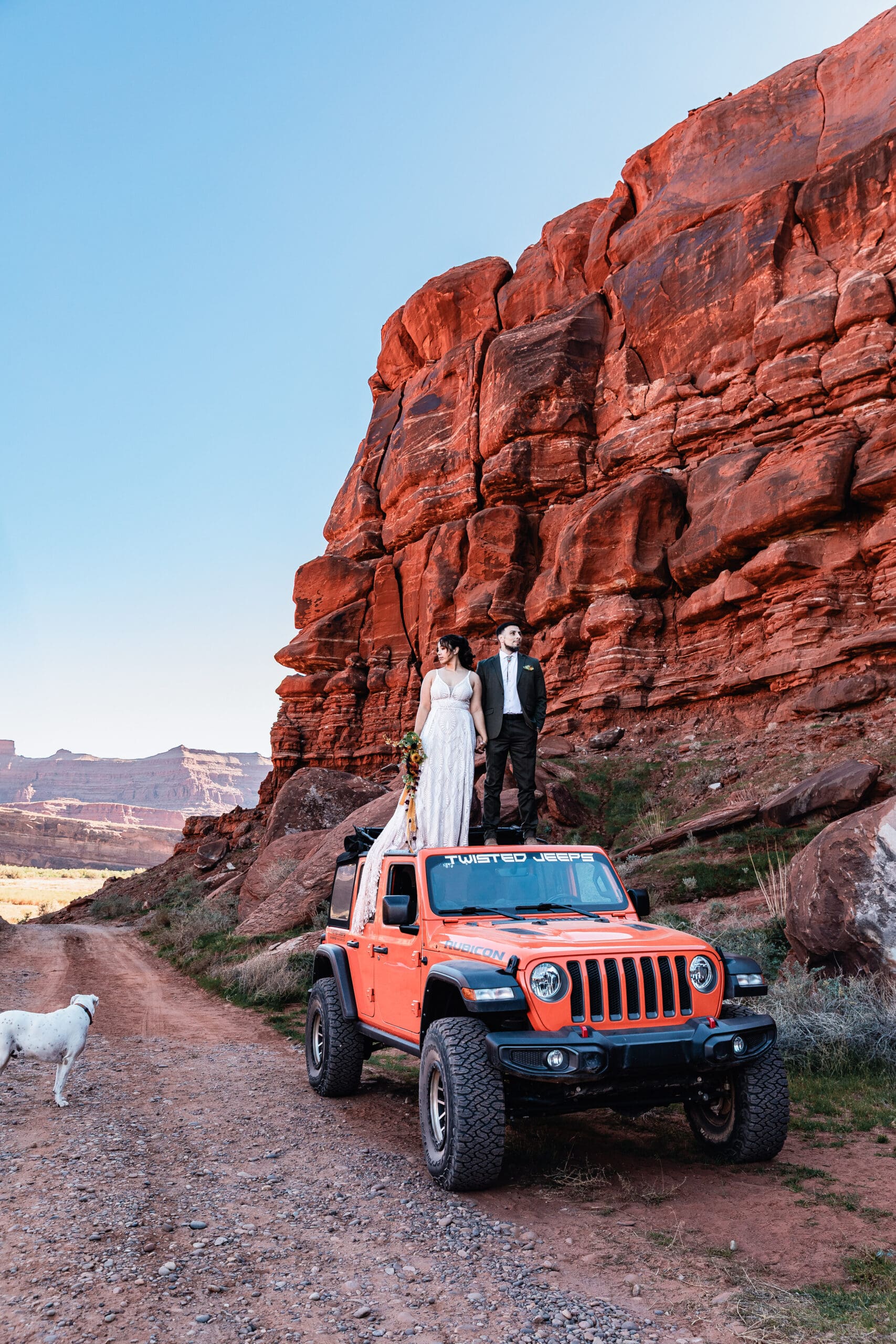 Couple standing on top of their Jeep during a Moab elopement with their dog looking up at them from the ground surrounded by red rock desert landscape